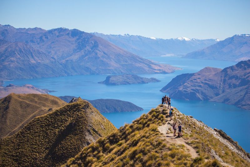 Roys Peak in Queenstown New Zealand.  There is a lake in the background, and there is a group of friends standing on top of the peak.
