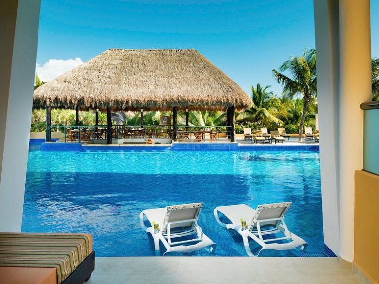 The gorgeous pool at El Dorado Seaside Palms. You can see the pool from the patio of a swim out suite. There are two white lounge chairs that look inviting in the water, and you can see the swim up bar beckoning from across the pool.